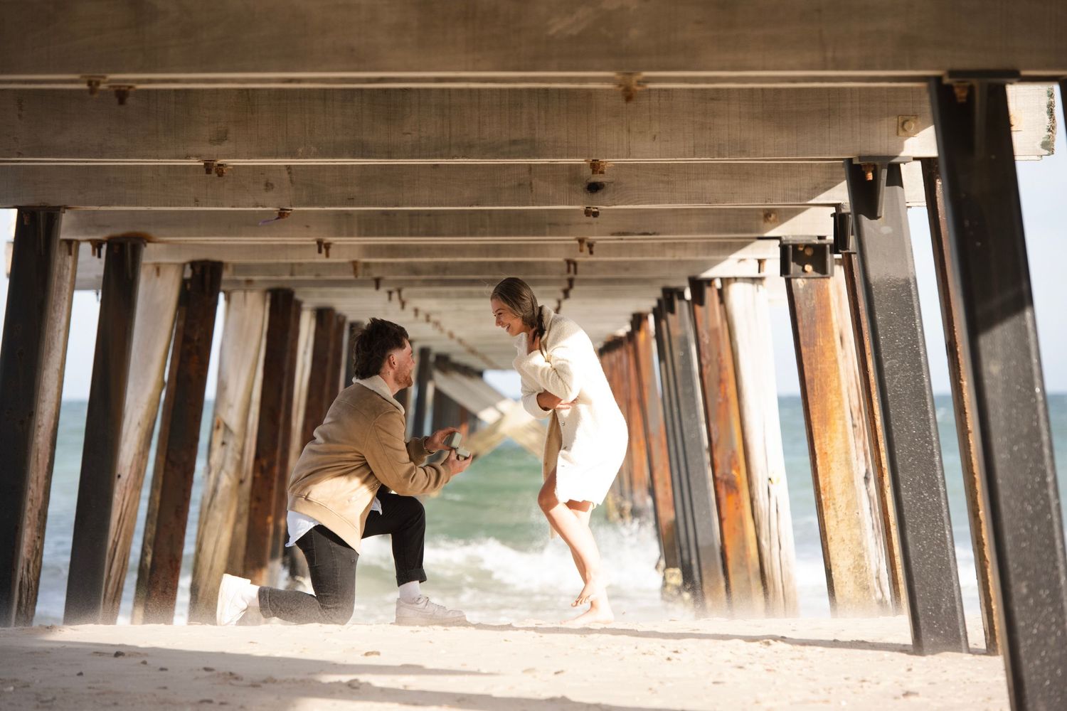 Man proposing on one knee to woman on sandy beach under wooden pier with ocean waves in background.