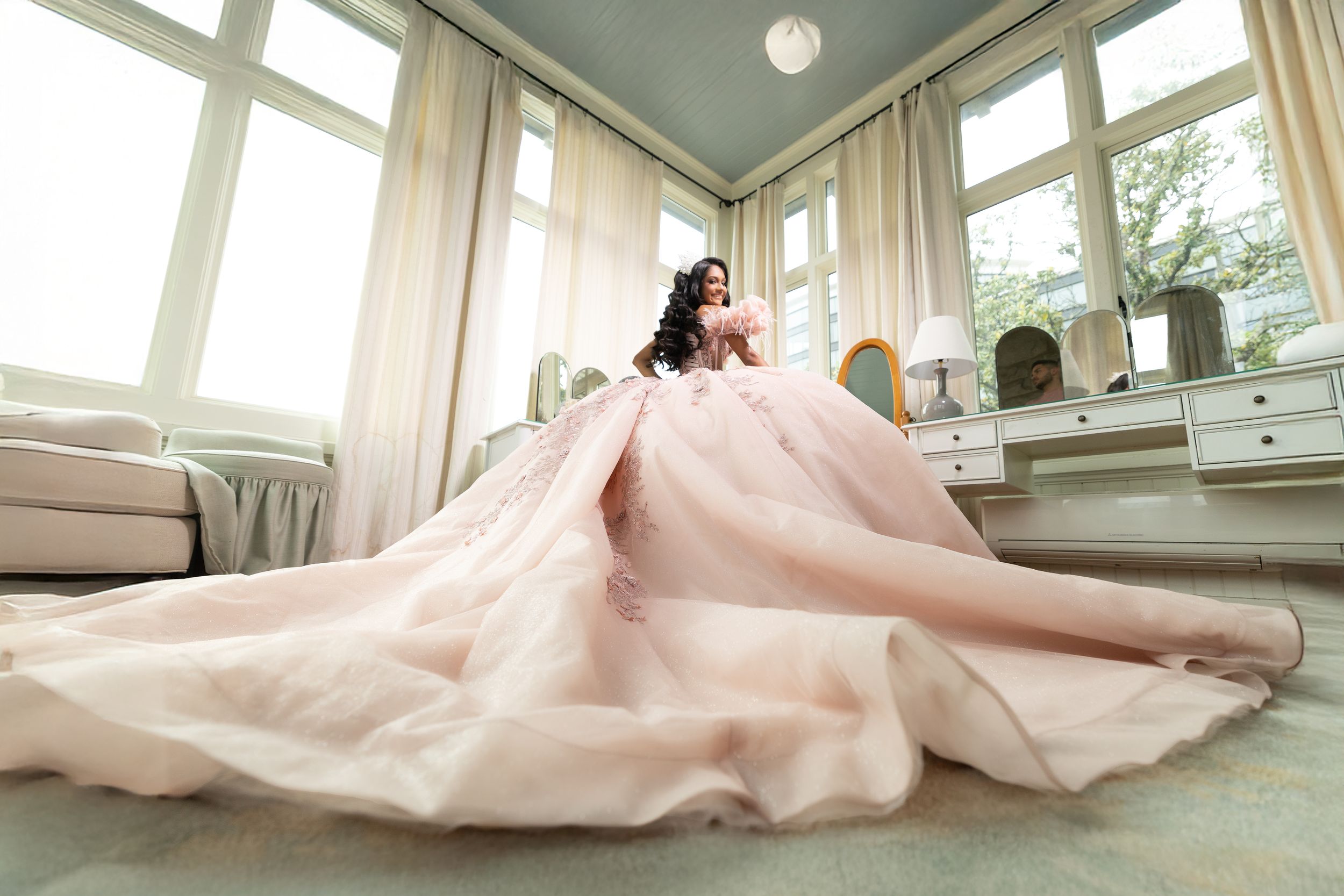 A flowing blush pink ballgown with a dramatic train fills an elegant sunlit room with white furniture and tall windows.