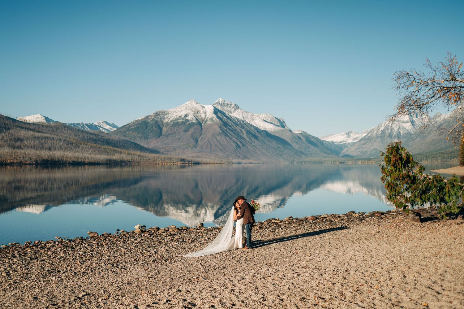 A scenic mountain lake with calm reflective waters surrounded by snow-capped peaks and rocky shoreline in Montana.