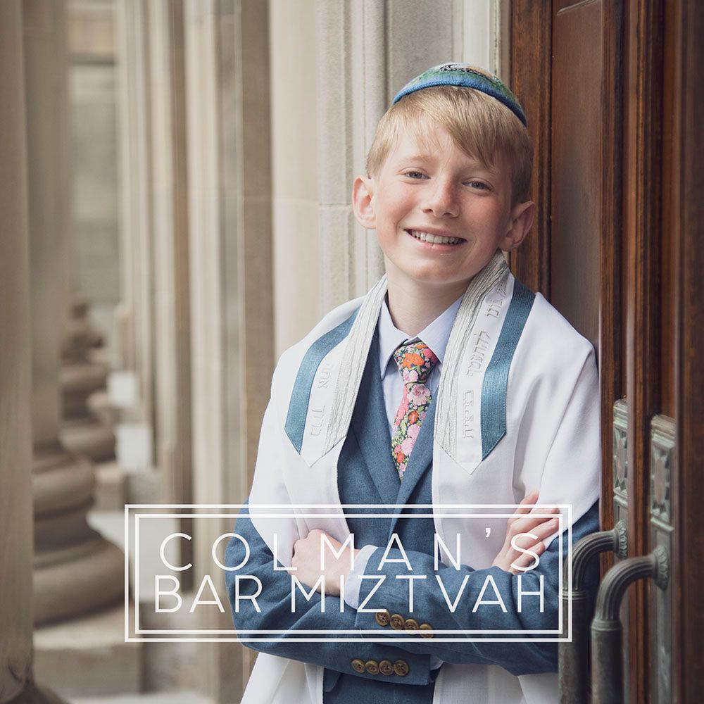 Young boy in a navy suit and blue tie smiles while standing in front of an elegant architectural backdrop.
