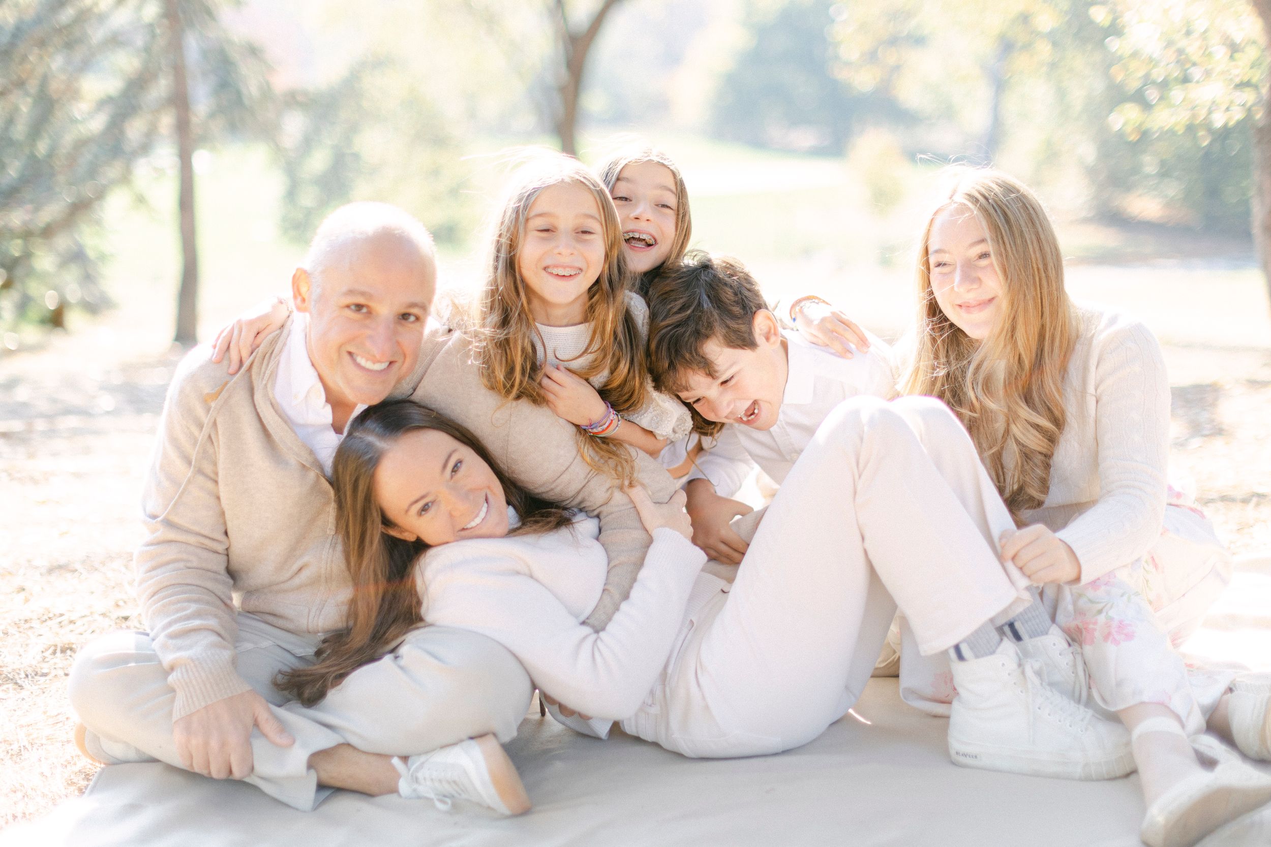 A family wearing white and beige clothing poses together on a light colored blanket during an outdoor photo session.