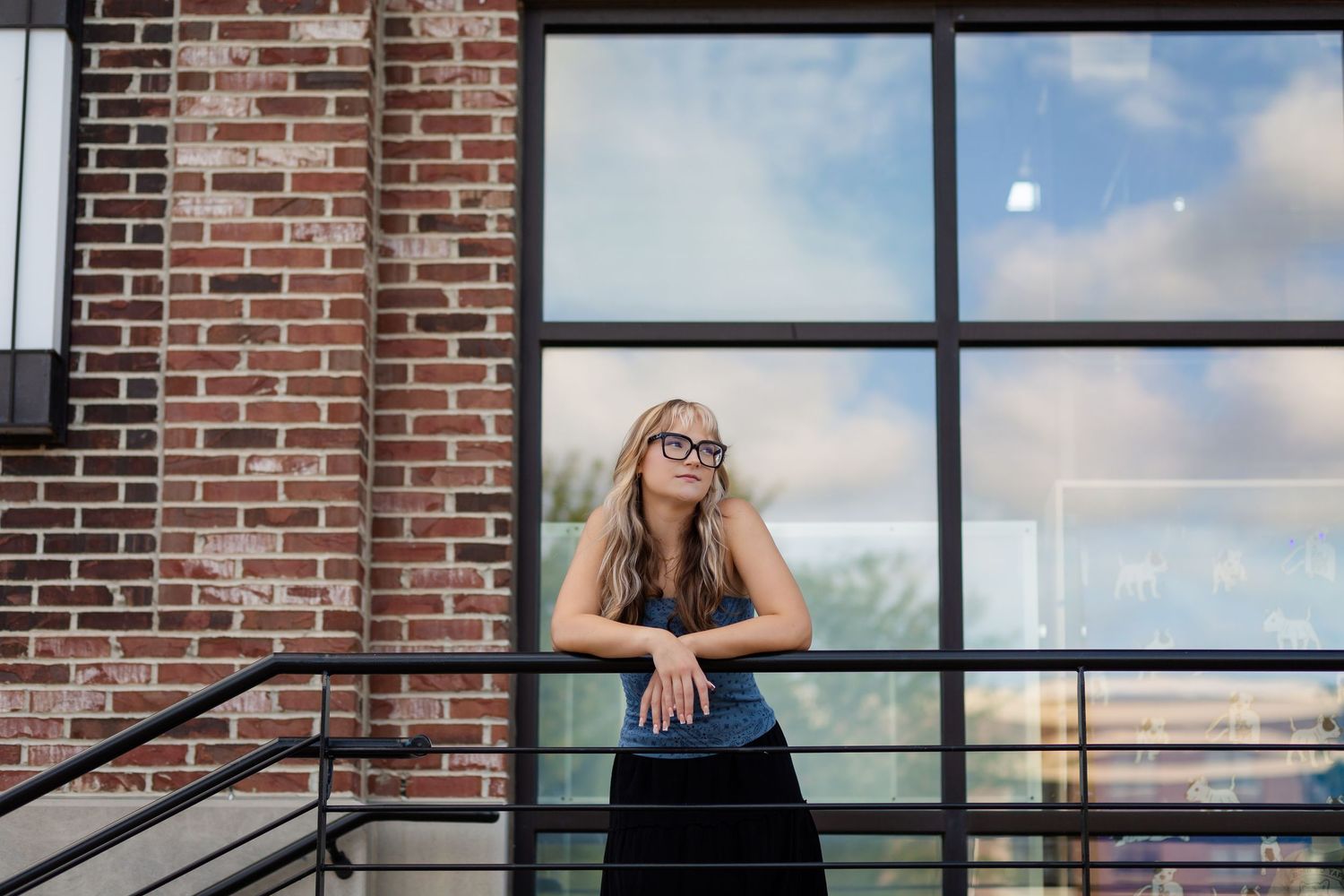 High school senior girl in glasses leans against a railing outside a modern brick building with large windows on a sunny day in NewBo.