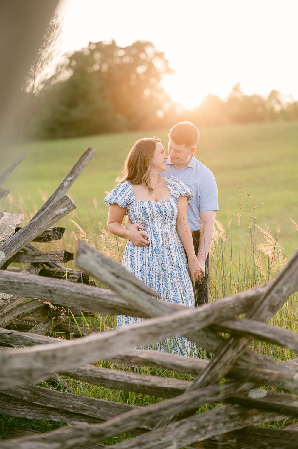 A couple embraces near wooden fence posts at sunset in a golden field with soft lighting.