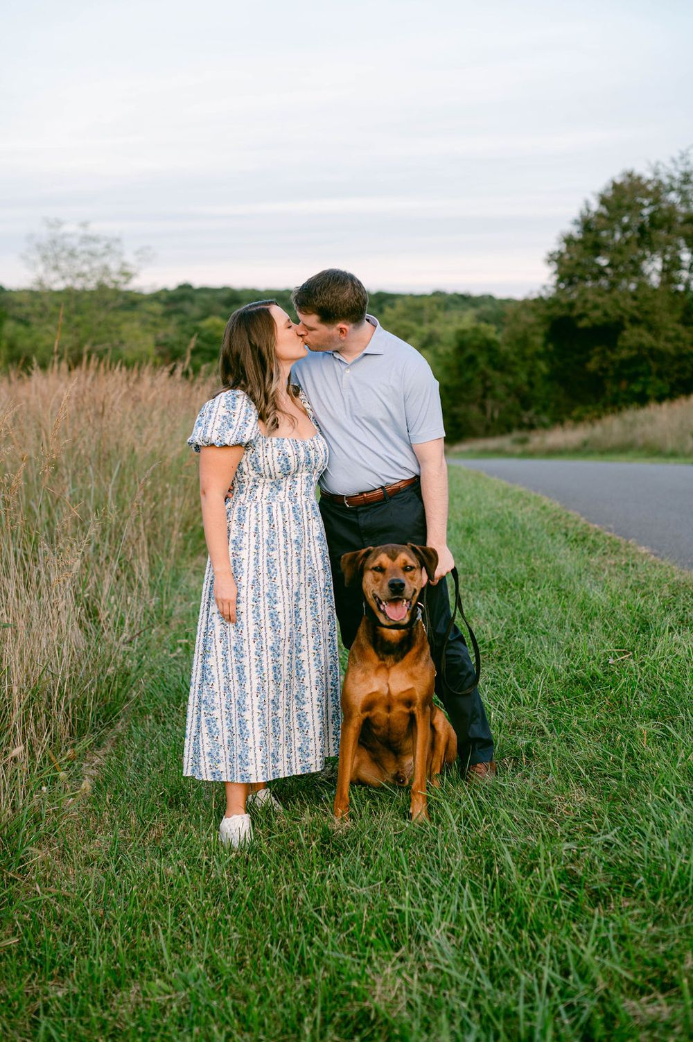 A couple poses with their brown dog in a grassy field during a sunset photoshoot.