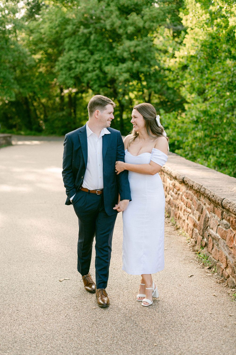 A couple strolls together on a stone-walled path surrounded by lush greenery on a sunny afternoon.