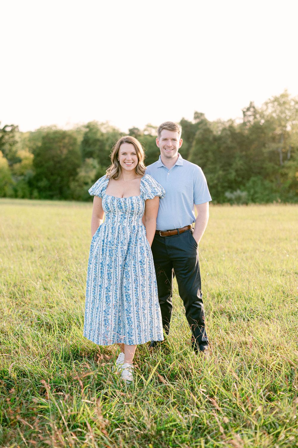 A couple stands together in a sunlit grassy field during a summer evening photoshoot.