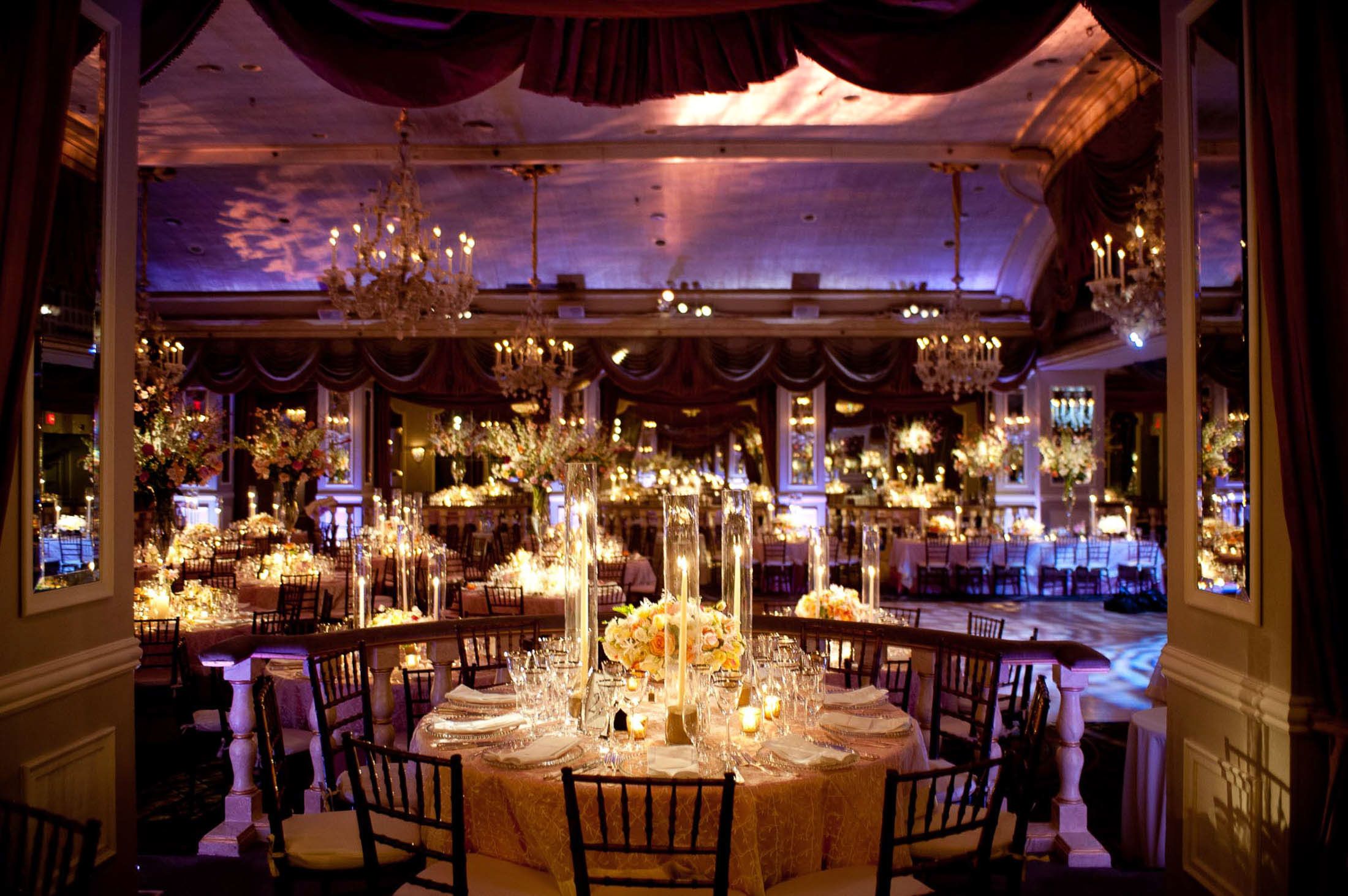 Elegant ballroom reception setup with crystal chandeliers, round tables, and dramatic purple lighting creating a romantic ambiance.