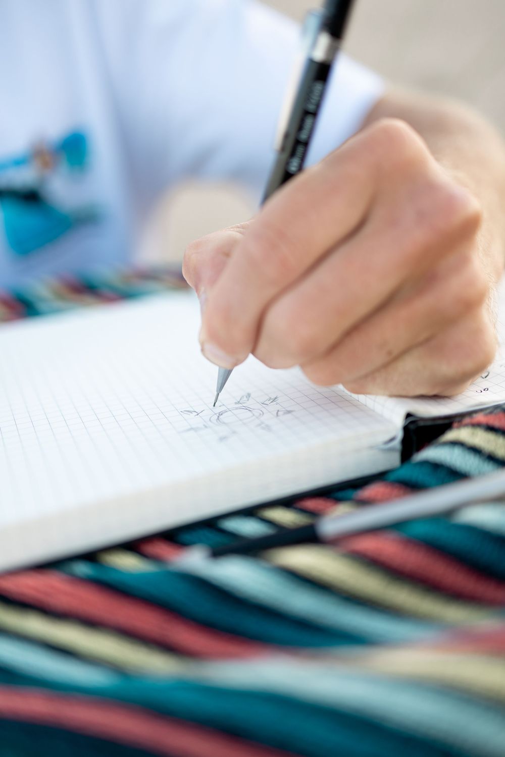 Close-up of a hand drawing floral sketches in a grid notebook with a black pen on a colorful striped surface.