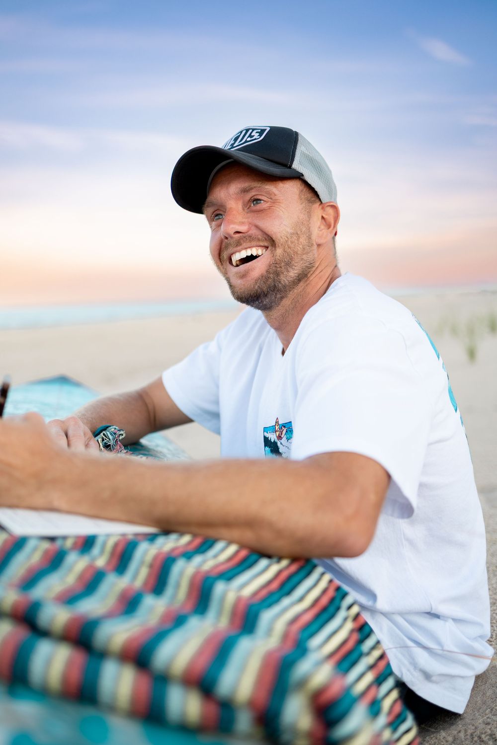 Smiling man in white t-shirt and baseball cap relaxing on a beach at sunset.