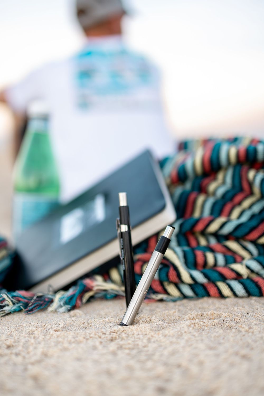 Two pens stuck in sand at beach with notebook, green bottle, and person relaxing on colorful blanket in background.