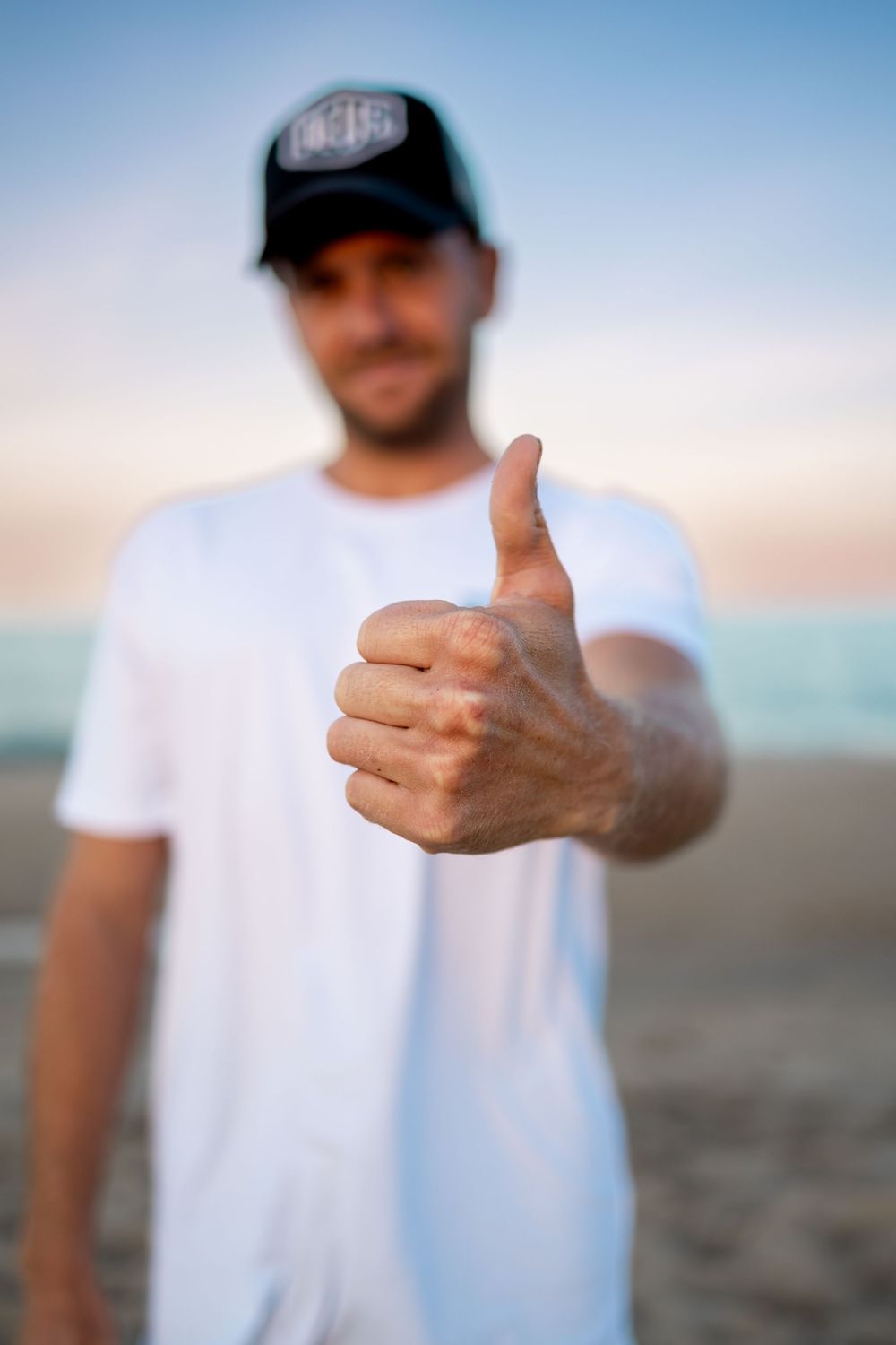 Man in white t-shirt and black cap giving thumbs up on beach at sunset.