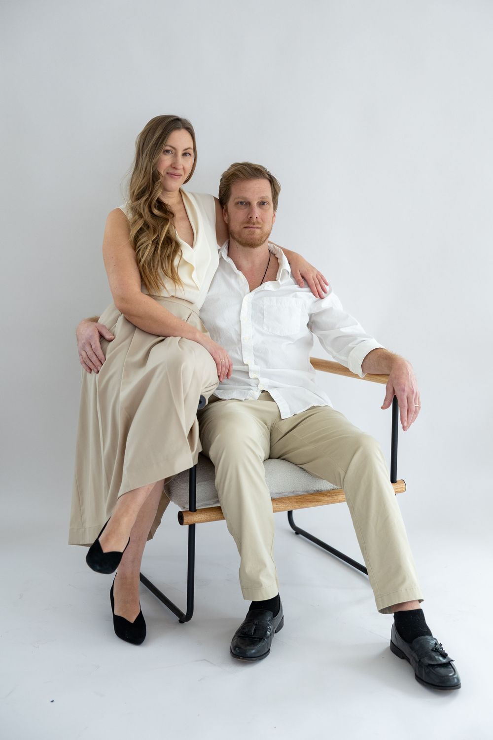 Two people in neutral colored outfits pose together on a chair against a white backdrop in a minimalist studio setting.