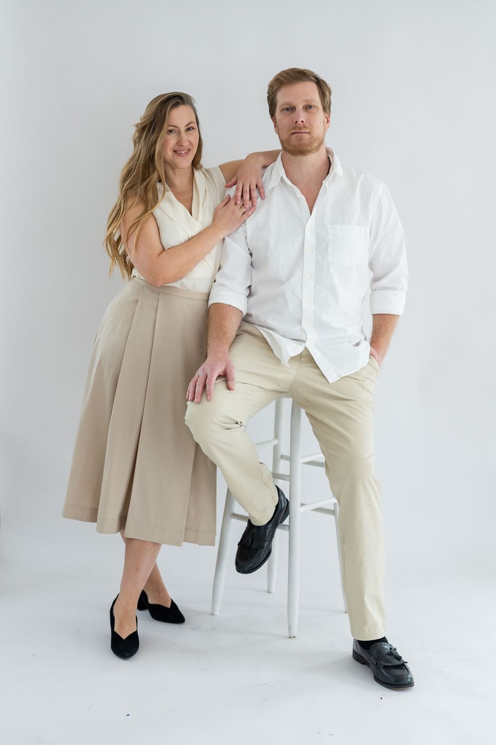 A couple in neutral-toned outfits poses together against a white backdrop in a studio setting.