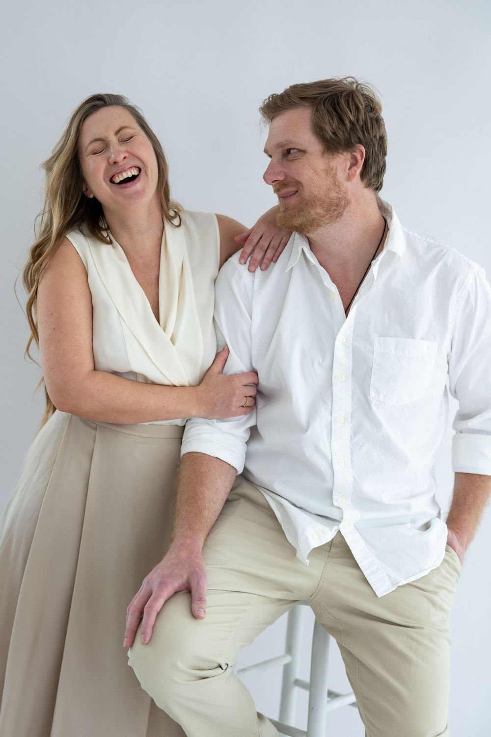A couple in white and beige clothing share playful moments together in a series of candid poses against a light background.