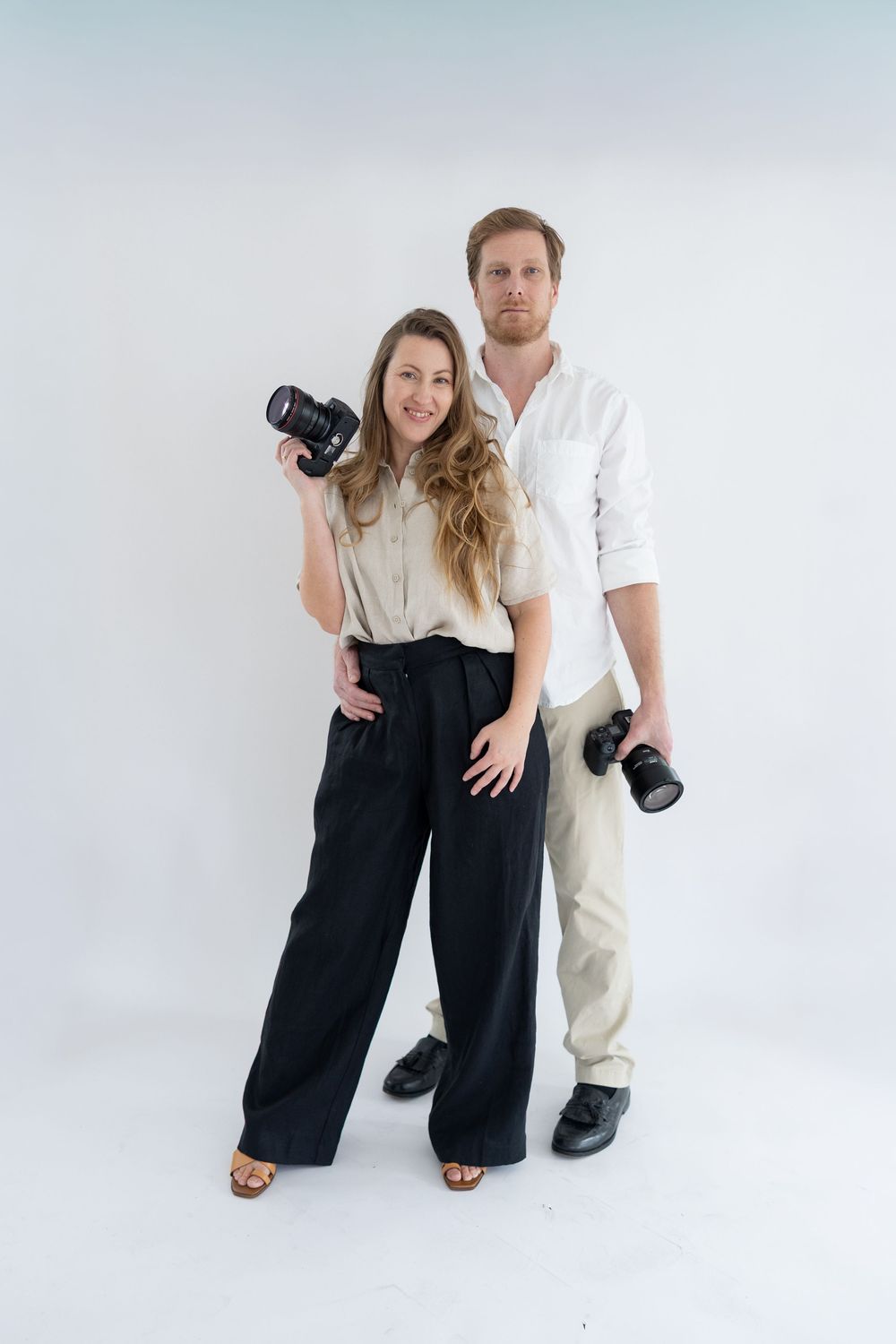 Professional photography duo posing together with camera equipment in a bright white studio setting.