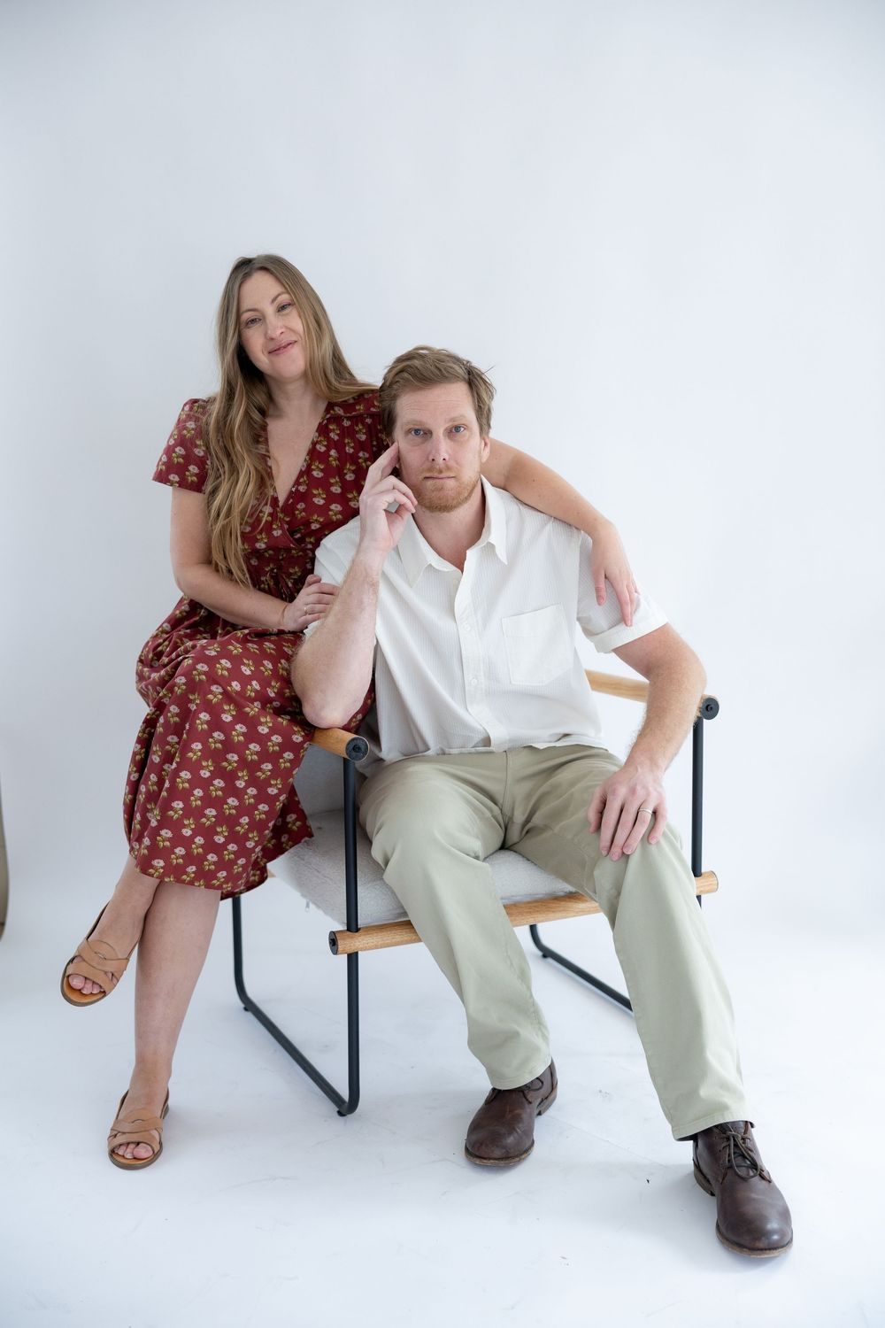 A couple poses together on a modern chair against a white backdrop in a studio setting.