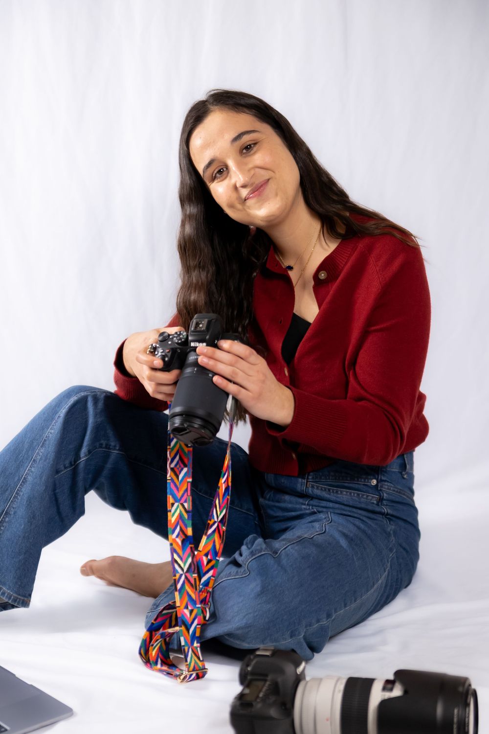 Photographer in red sweater and jeans reclines while holding camera and reviewing photos on white backdrop.