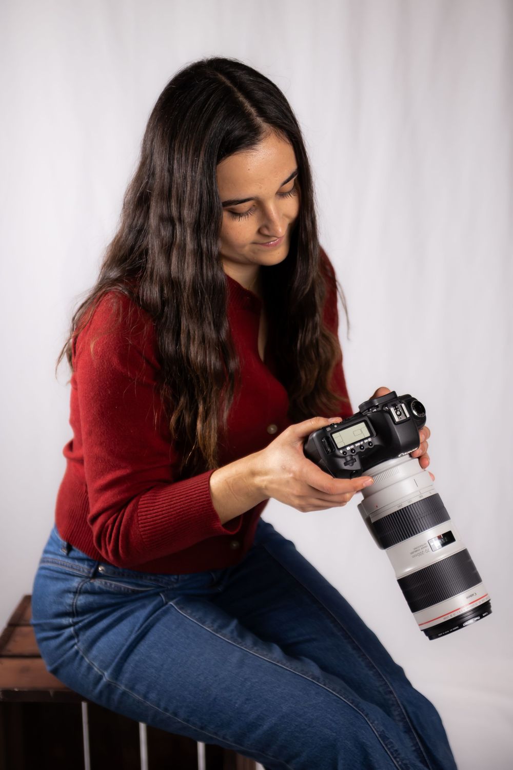 A photographer in a red sweater and jeans examines settings on a professional camera with telephoto lens.