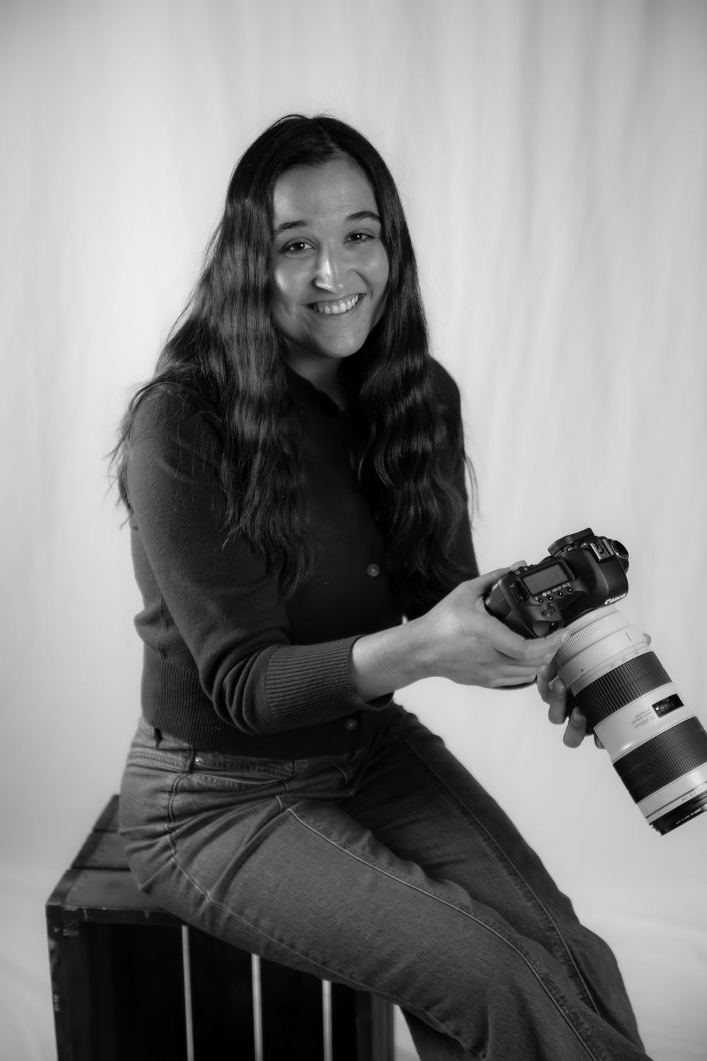 Black and white portrait of photographer sitting with professional camera and telephoto lens against light background.