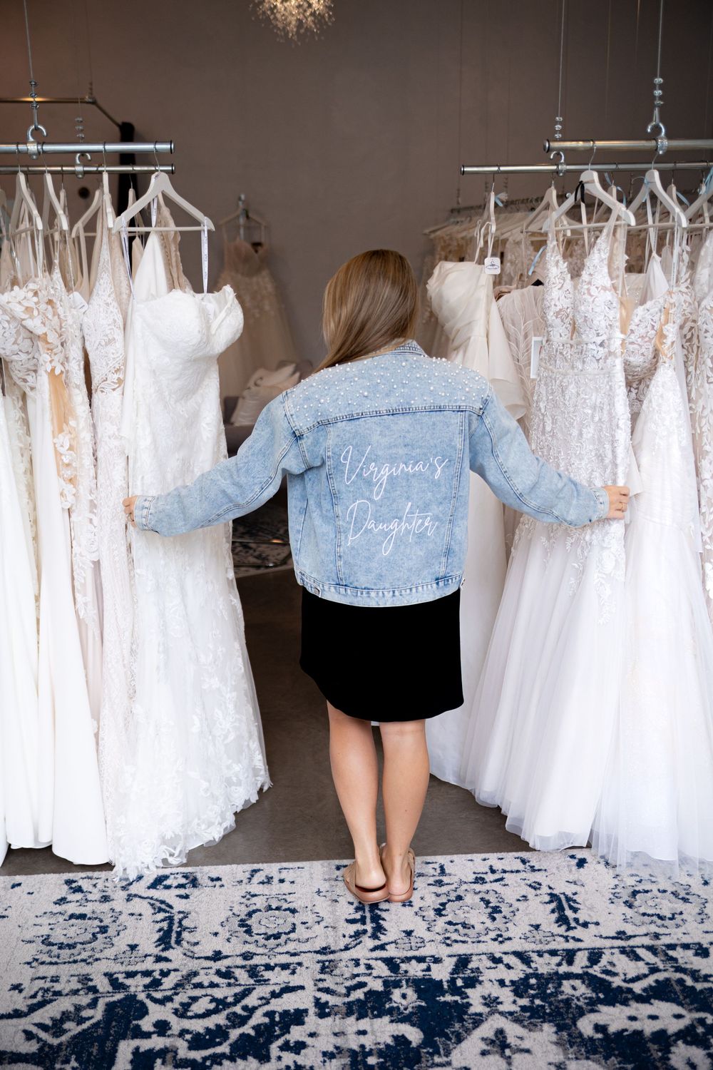 A series of white wedding dresses hanging on a rack against a dark wall with a blue patterned rug below.