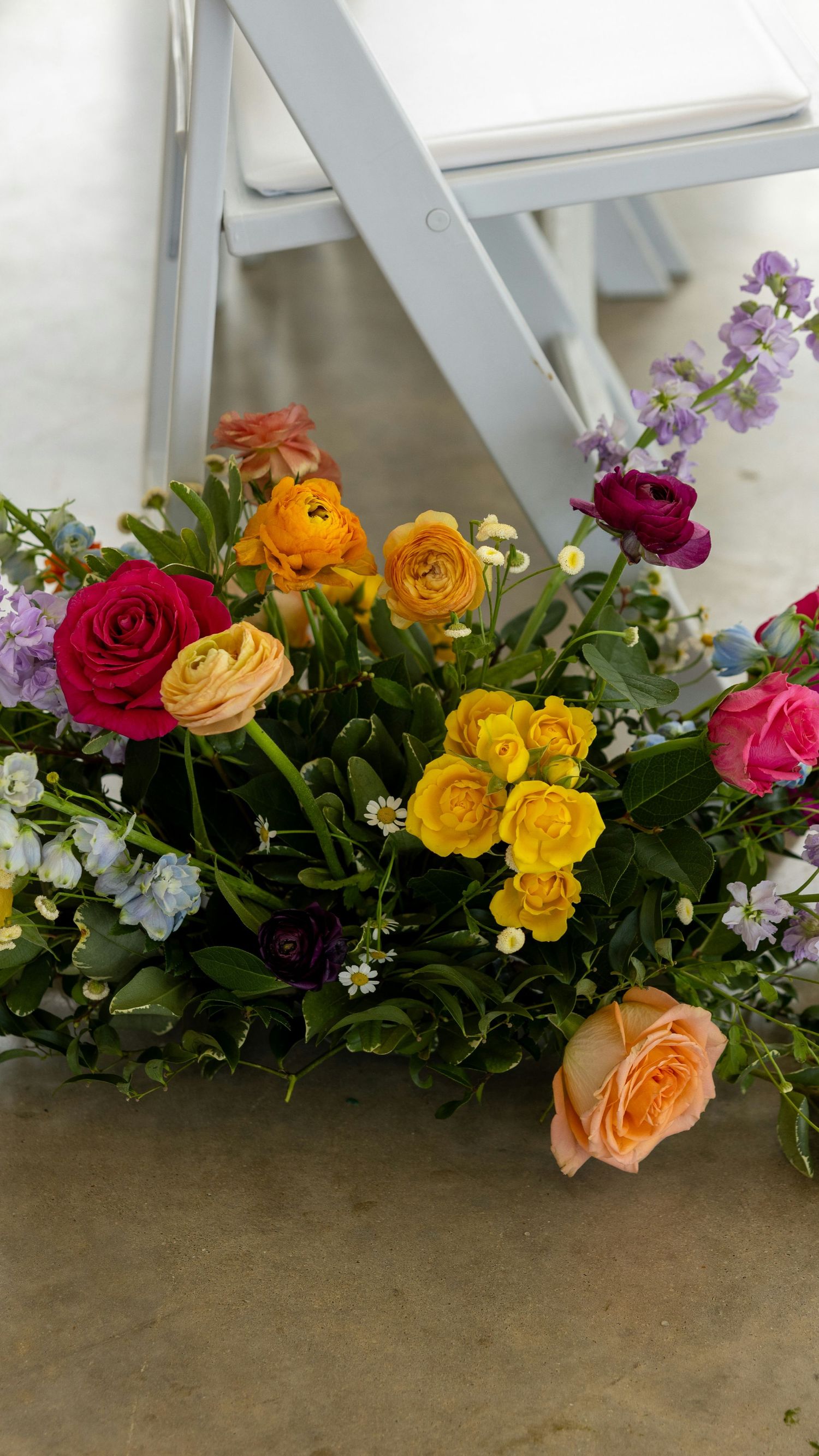 Vibrant wedding floral arrangement with roses and ranunculus in rainbow colors beside a white chair.