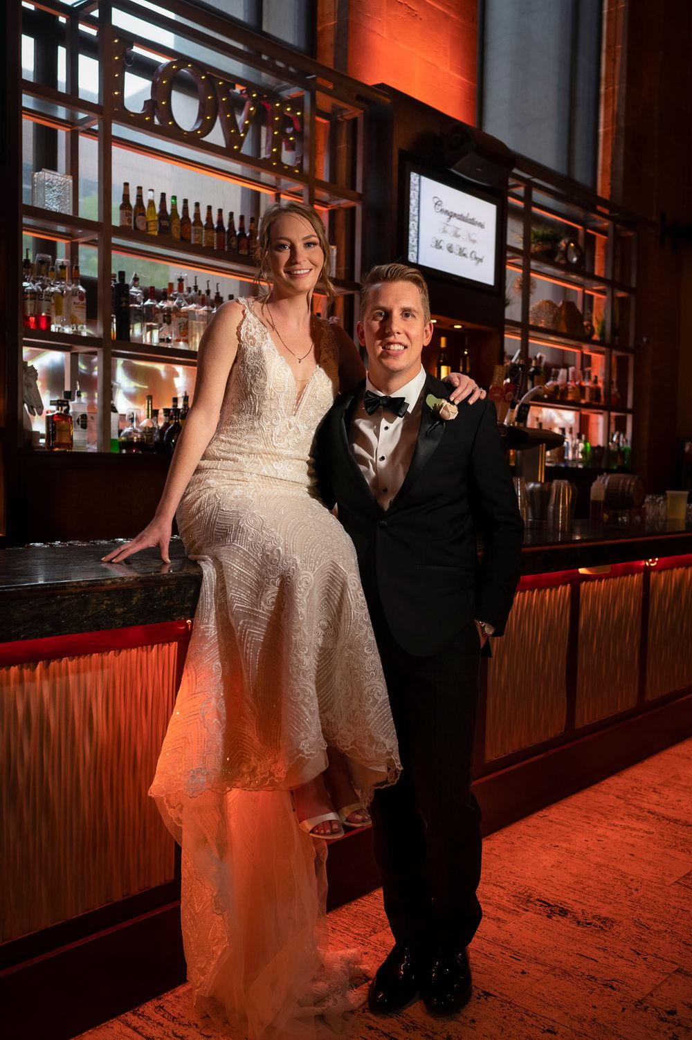 A couple in formal attire poses together at a dimly lit bar with warm orange lighting at Union Trust in Philadelphia.