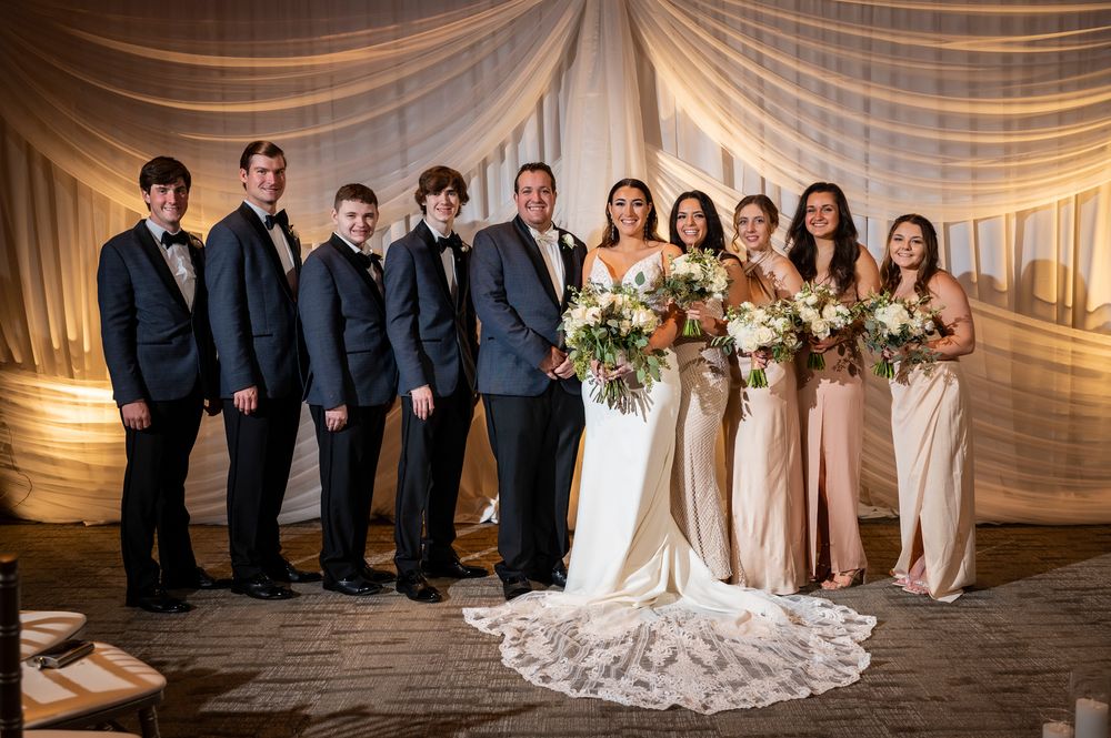 Wedding party poses together in formal attire against elegant draped fabric backdrop in a reception venue.