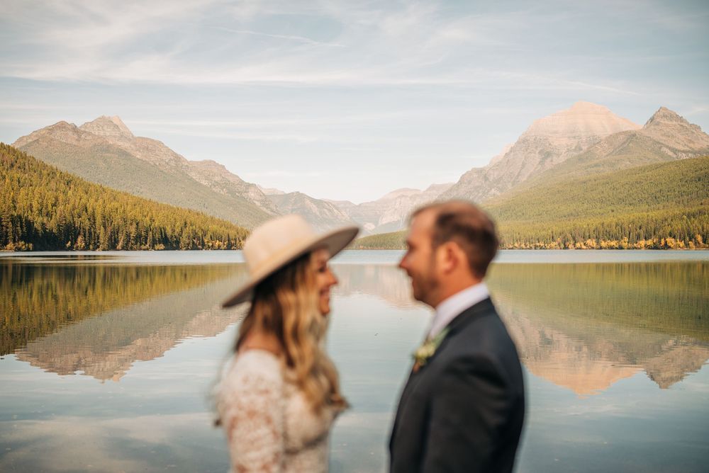 A romantic couple embraces by a serene mountain lake with snow-capped peaks and evergreen forests in the background.
