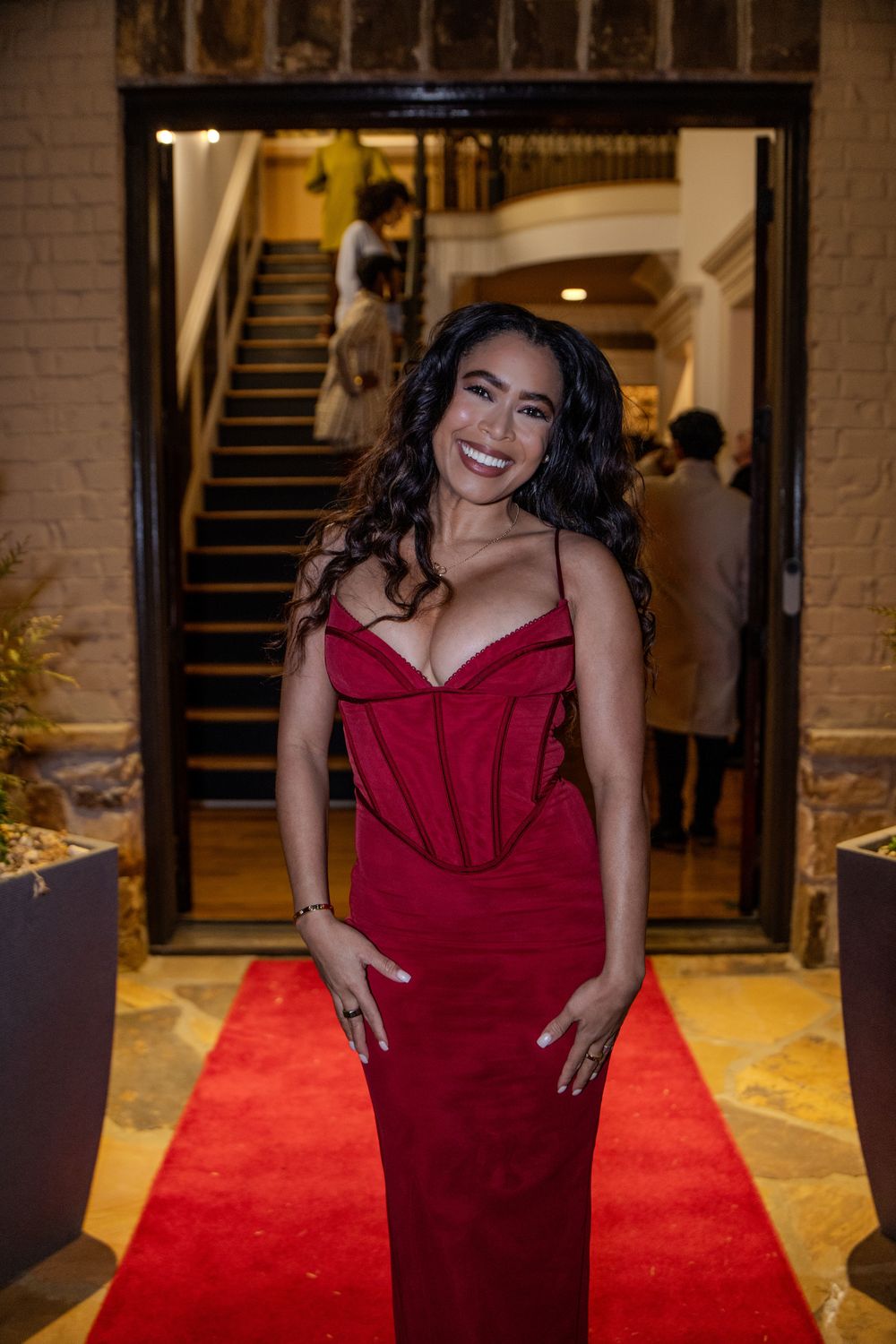 Woman in a burgundy formal dress poses on a red carpet in front of a staircase with warm lighting.
