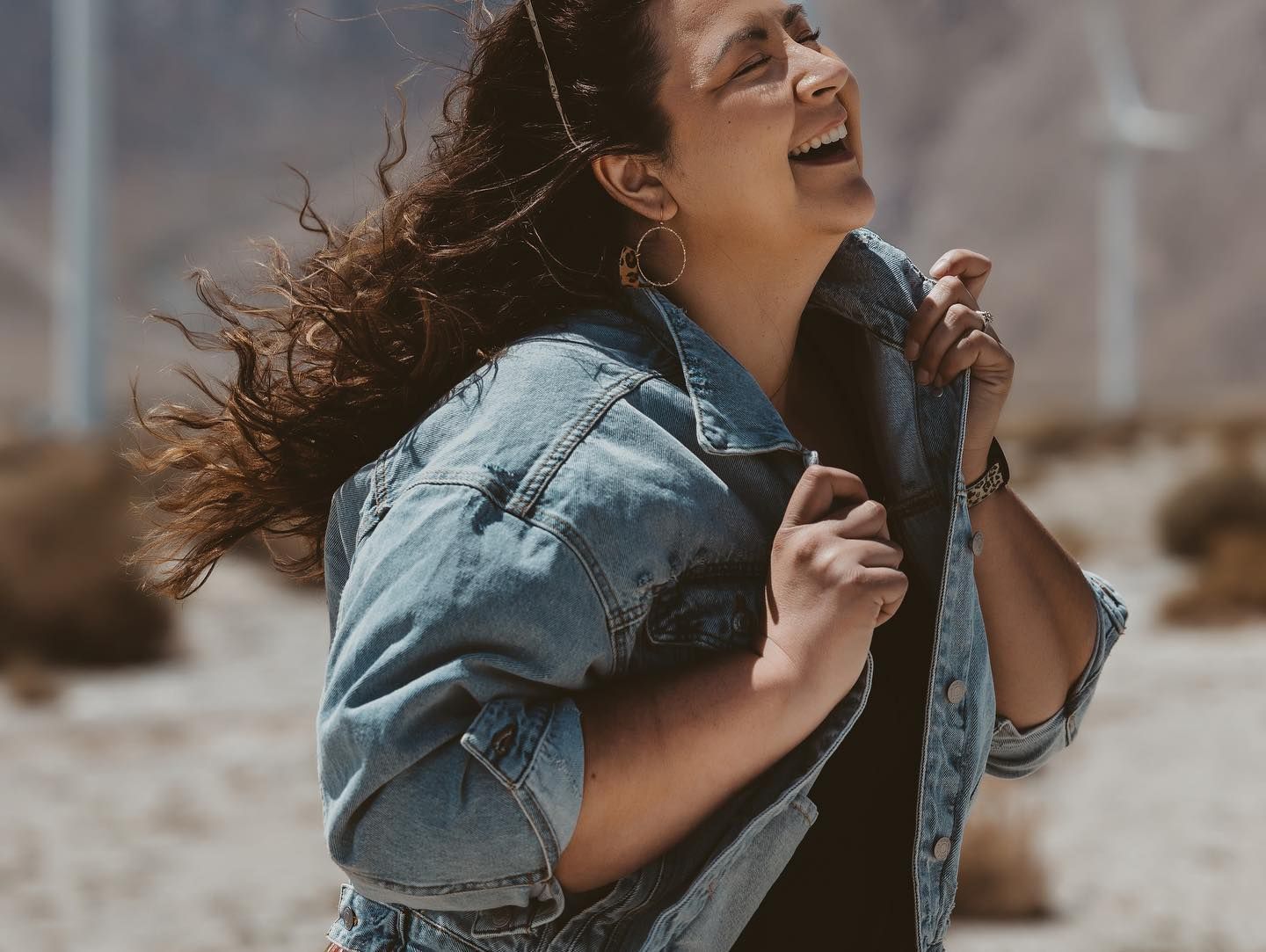 Woman in denim jacket laughing with wind-blown hair in desert landscape.