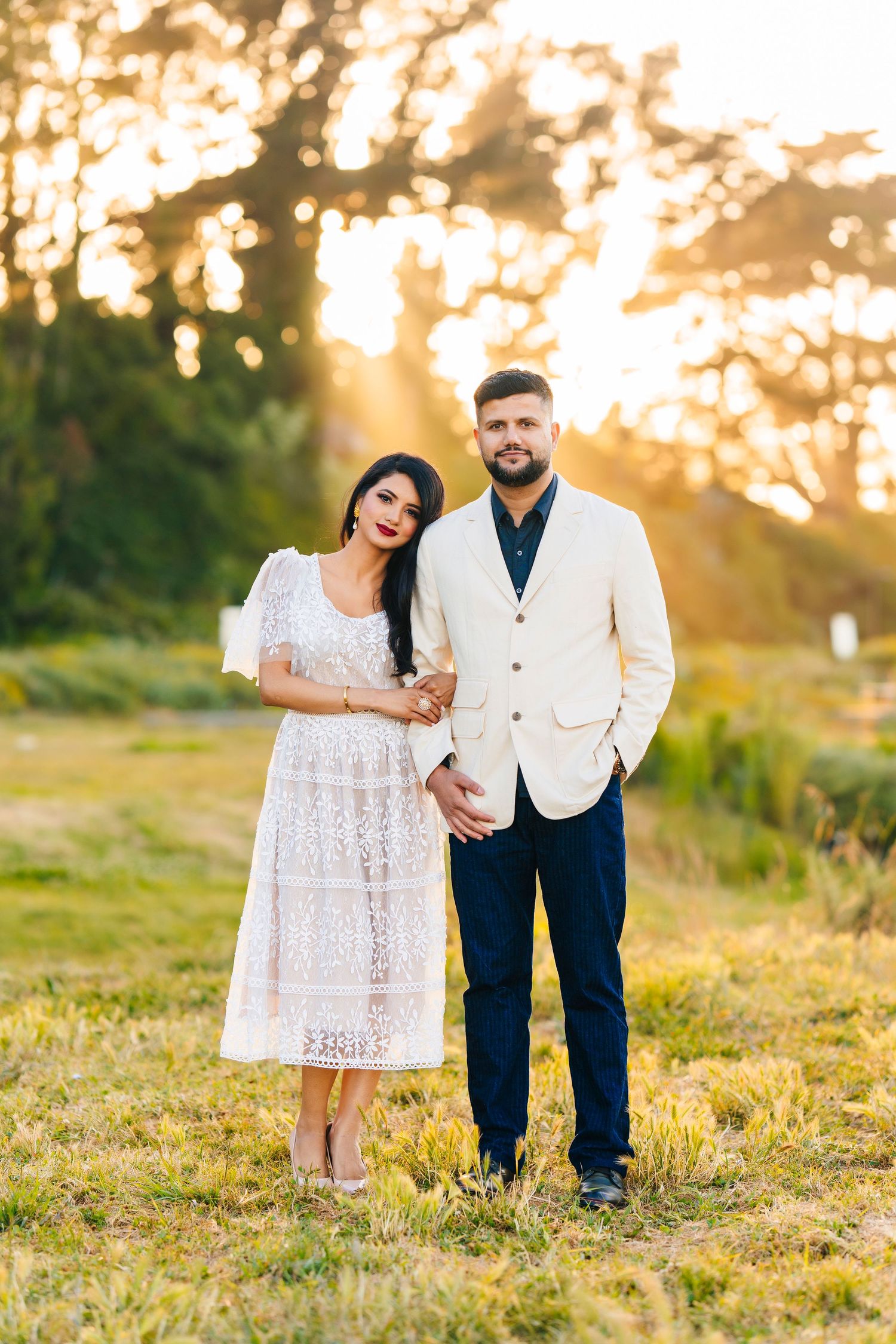 A couple stands together in formal wear during sunset in a natural outdoor setting.