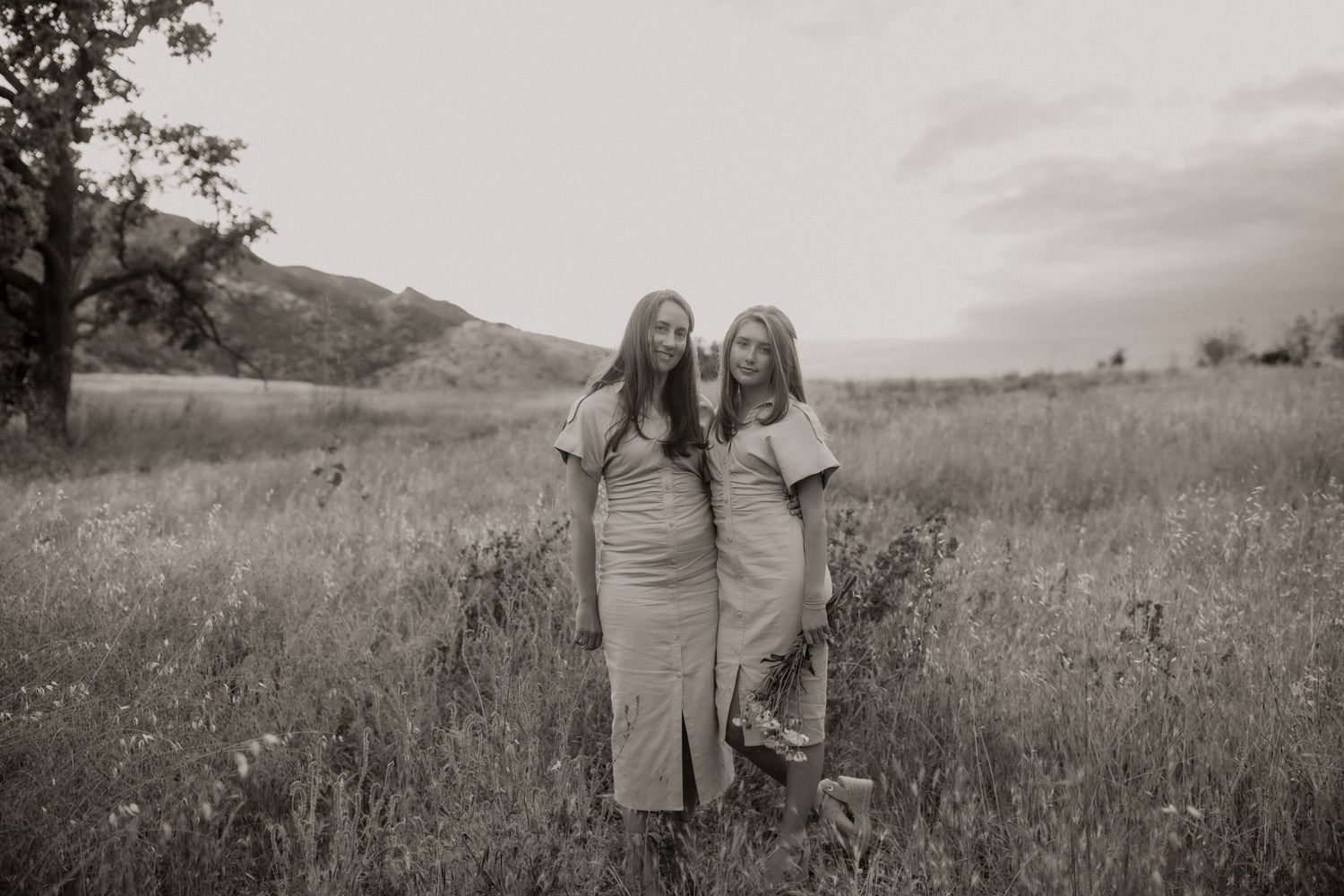 Two people stand together in a meadow with mountains in the background in black and white photography.