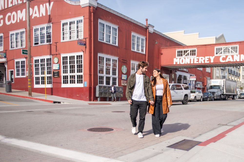 A couple walks together past a red industrial building on a city street corner.