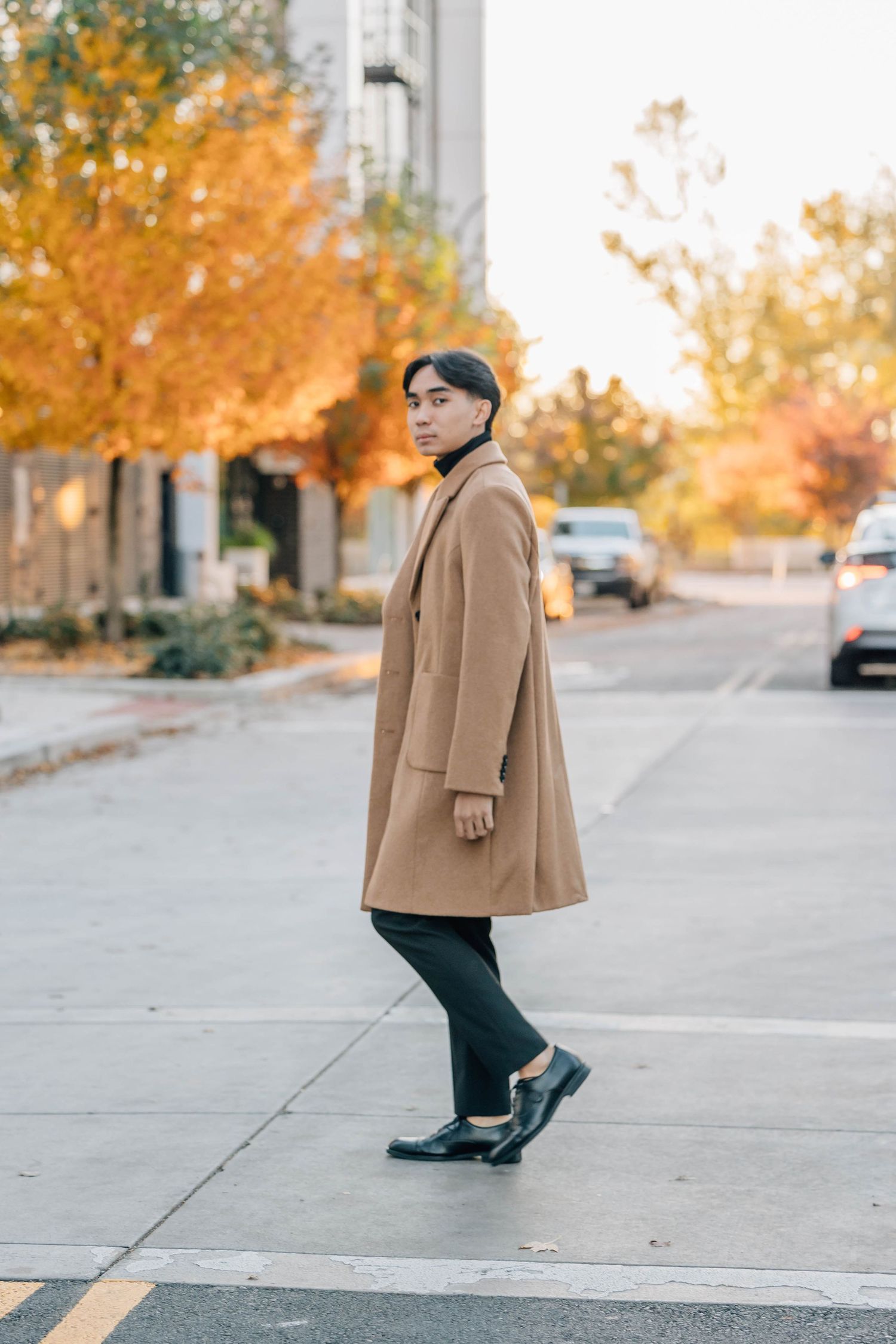 Young Asian man in camel overcoat crossing street with autumn foliage trees in background.