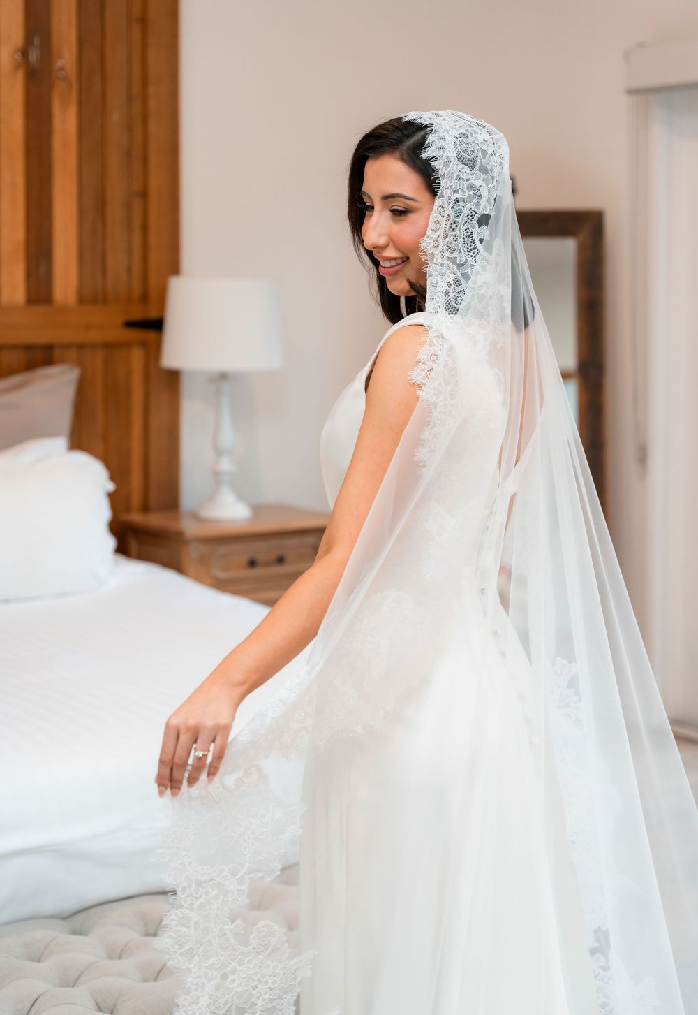 A bride in a white wedding gown and long veil sits elegantly on a tufted ottoman in a bridal suite.