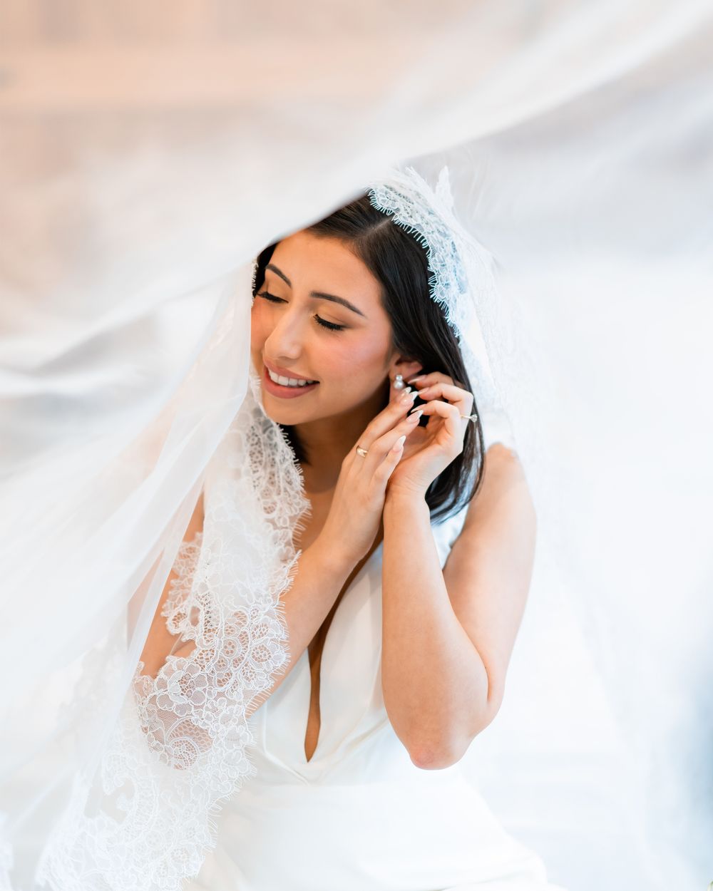 Bride in delicate white lace veil poses gracefully in ethereal bridal portraits against soft light background.
