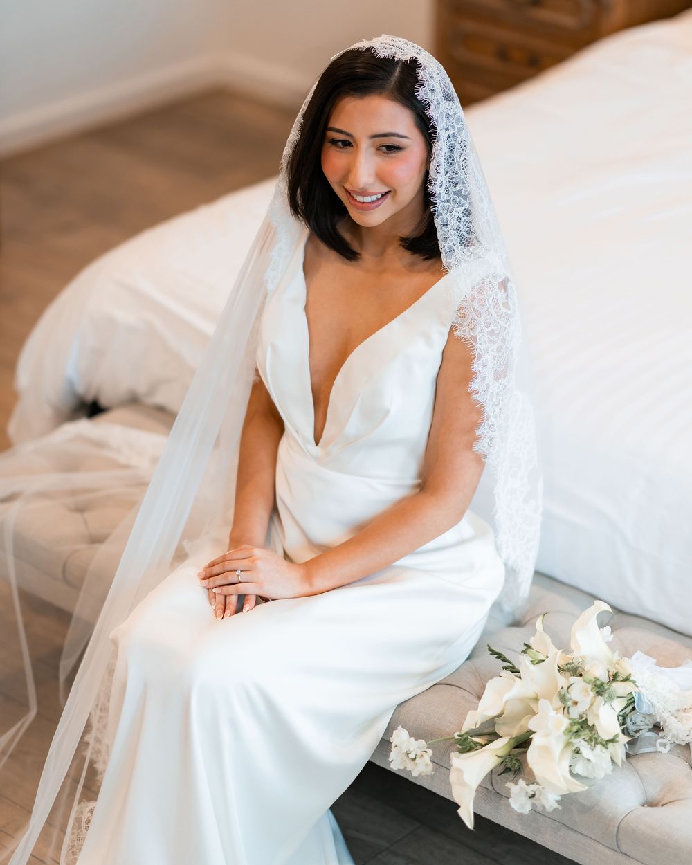 A bride in a white wedding dress sits elegantly on a couch with a delicate veil and white floral bouquet.
