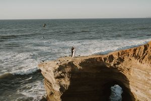 A lone figure stands on a dramatic ocean cliff arch formation at sunset with crashing waves below.