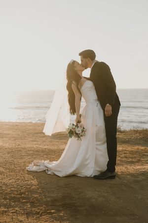 A romantic wedding couple shares a kiss at sunset on a coastal cliff overlooking the ocean.