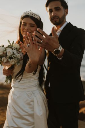 Happy newlyweds showing off their wedding rings with a bridal bouquet and sunset backdrop.
