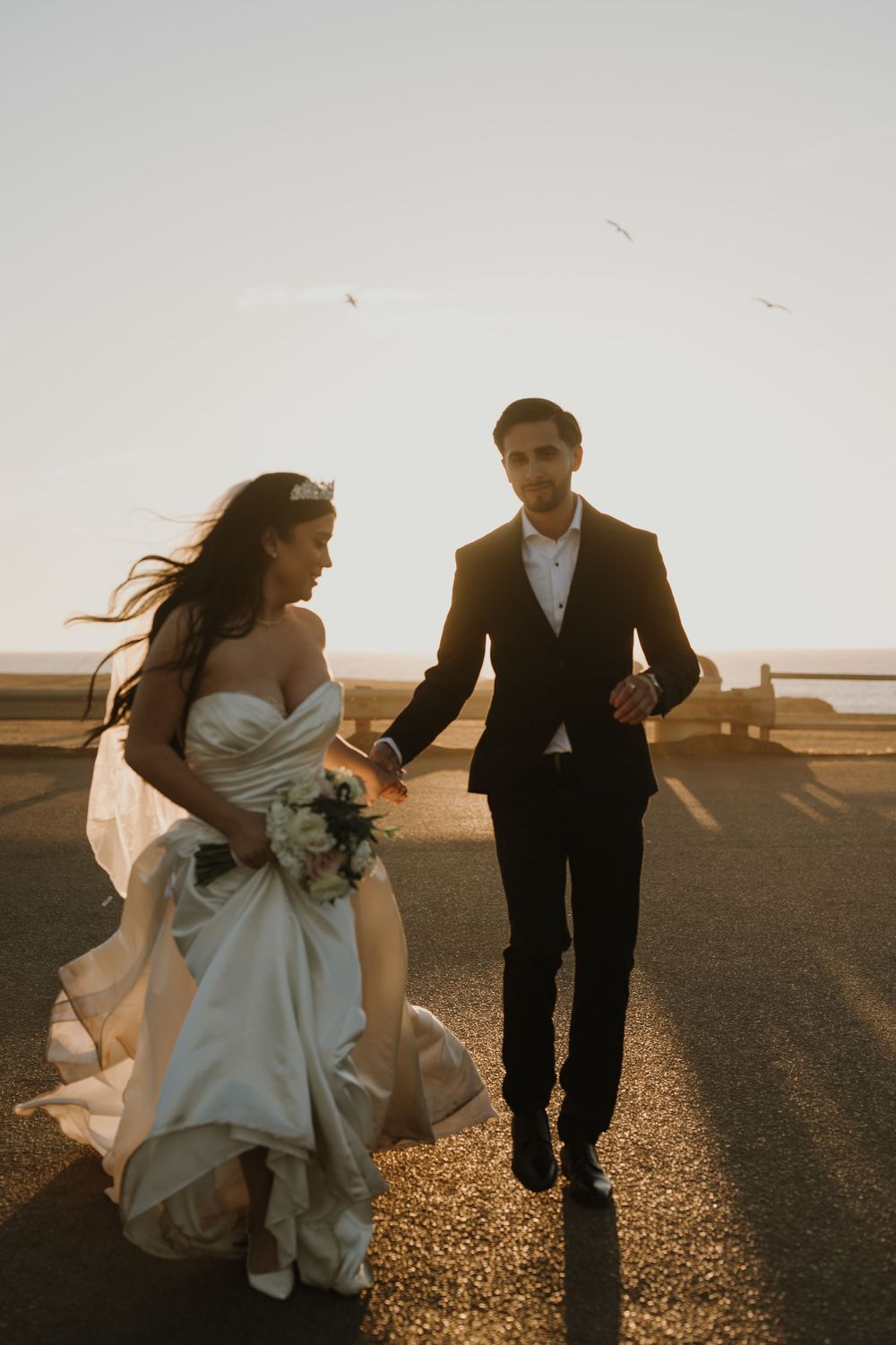 Newlyweds walk hand in hand along a coastal path at golden hour with flowing dress and veil.