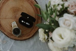 Wedding details with rings, watch and flowers arranged on a rustic wooden slice.
