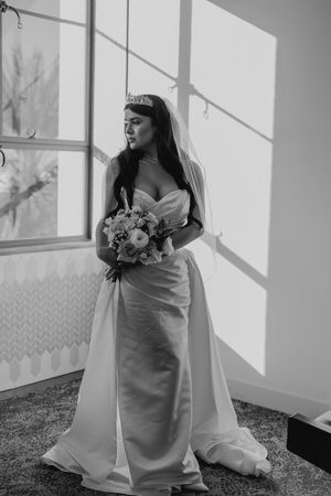 Bride in classic white gown poses by window with bouquet in black and white portrait.