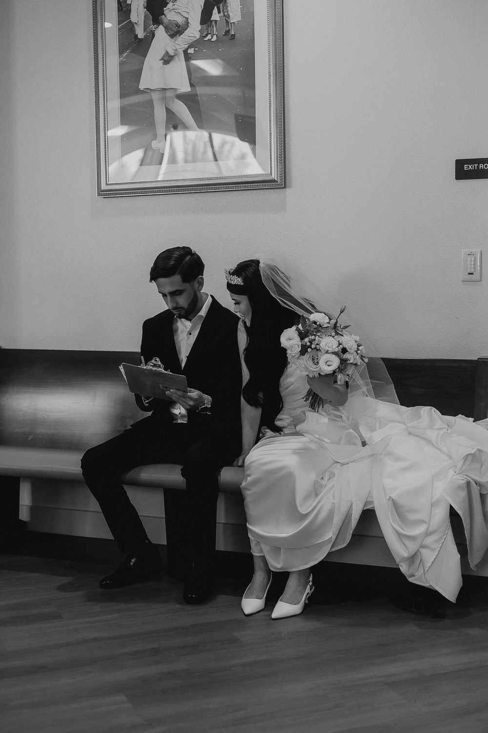 Wedding couple sits on bench looking at document beneath framed photograph on wall.
