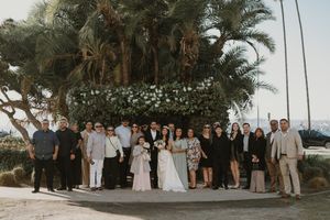 Wedding party and guests gathered outdoors among palm trees for a group photo.