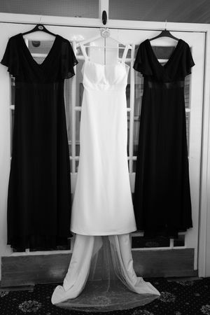 A white wedding gown hanging between two black bridesmaid dresses on display against a white door in black and white.