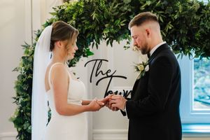 A couple exchanges rings during their wedding ceremony under a lush green floral arch.