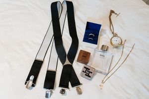 Wedding accessories including black suspenders, rings, jewelry and cologne bottles arranged on white fabric.