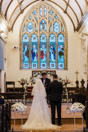 Wedding ceremony inside historic church with beautiful blue stained glass window and white floral arrangements at altar.
