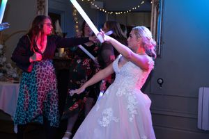 Person wearing a white gown strikes a dramatic lightsaber pose under colorful lighting at a dance party.